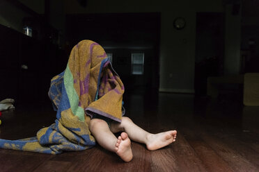 Verspielter kleiner Junge, der seinen Kopf mit einem Handtuch bedeckt, während er zu Hause auf dem Boden sitzt - CAVF49425