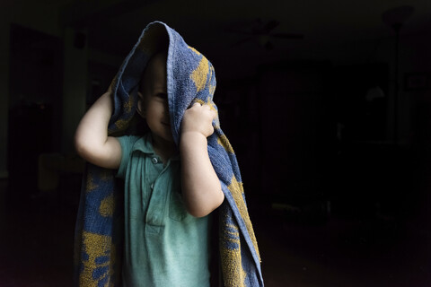 Porträt eines niedlichen kleinen Jungen, der mit einem Handtuch spielt, während er in der Dunkelkammer zu Hause steht, lizenzfreies Stockfoto