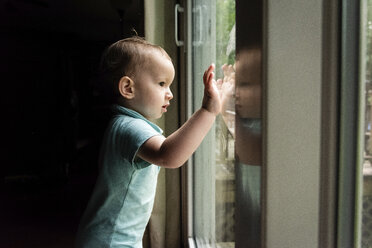 Side view of cute baby boy looking through window while standing in darkroom at home - CAVF49427