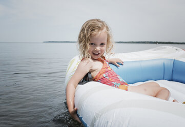 Porträt eines niedlichen Mädchens, das in einem aufblasbaren Floß auf einem See gegen den Himmel sitzt - CAVF49431