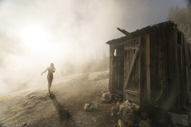 Silhouette einer Frau auf dem Weg zu einer alten Holzhütte auf einem Berg bei nebligem Wetter - CAVF49523