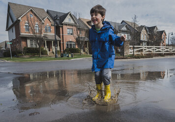 Full length of playful boy wearing rubber boots while jumping in puddle on road against houses - CAVF49525