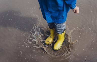 Low section of playful boy wearing yellow rubber boots while splashing puddle - CAVF49526