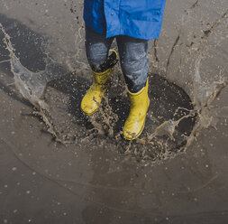 Low section of playful boy wearing rubber boots while splashing puddle - CAVF49527