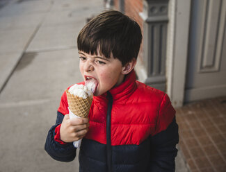 Boy looking away while licking ice cream on footpath in city - CAVF49528
