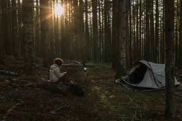 Woman using mobile phone while sitting at campsite in forest during sunset - CAVF49545