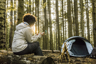 Woman using mobile phone while sitting on log at campsite in forest - CAVF49546