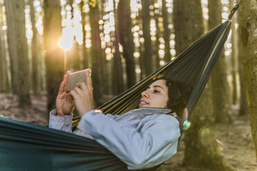 Frau, die ein Mobiltelefon benutzt, während sie bei Sonnenuntergang in einer Hängematte im Wald liegt - CAVF49547