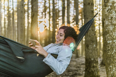 Frau, die ein Smartphone benutzt, während sie bei Sonnenuntergang in einer Hängematte im Wald liegt - CAVF49548