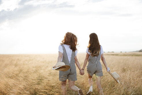 Rear view of happy twin sisters holding hands while walking on grassy field against sky during sunset - CAVF49610