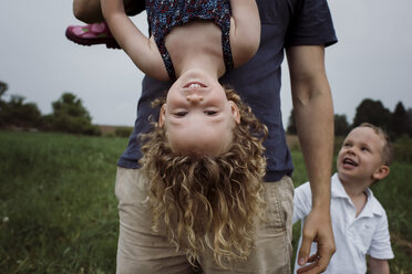 Mittelteil eines Vaters, der seine Tochter auf den Kopf stellt, während er mit seinem Sohn auf einer Wiese vor dem Himmel im Park steht - CAVF49617