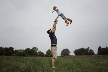 Happy father throwing son in air while standing on grassy field against sky at park - CAVF49618