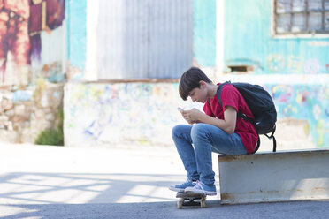 Seitenansicht eines Studenten mit Skateboard, der ein Mobiltelefon benutzt, während er in der Stadt sitzt - CAVF49627