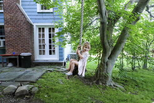 Nettes glückliches Mädchen, das wegschaut, während es auf einer Seilschaukel im Garten schwingt - CAVF49637