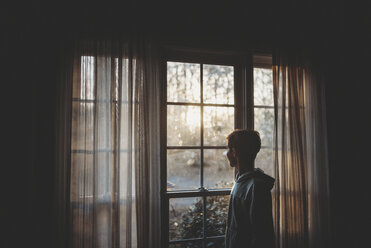 Side view of teenage boy looking through window while standing at home - CAVF49650