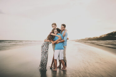 Happy siblings embracing while standing at beach against sky during sunset - CAVF49655
