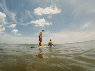 Shirtless brothers paddleboarding on sea against sky - CAVF49658