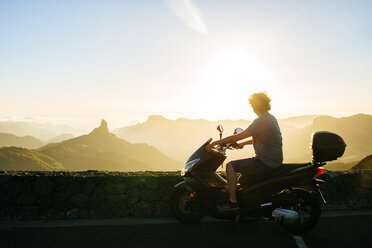 Spanien, Kanarische Inseln, Gran Canaria, Mann auf Motorroller beobachtet Sonnenuntergang über Berglandschaft - KIJF02064