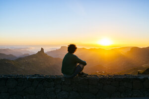 Spanien, Kanarische Inseln, Gran Canaria, Rückenansicht eines Mannes, der auf einer Mauer sitzt und den Sonnenuntergang über einer Berglandschaft beobachtet - KIJF02067