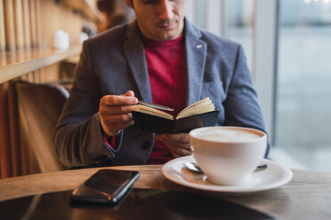 Businessman sitting in restaurant drinking coffee and checking his notebook - KKAF02439