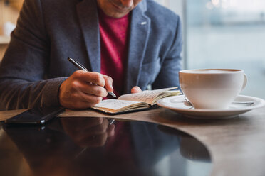 Businessman sitting in restaurant drinking coffee and checking his notebook - KKAF02441