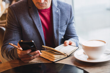 Businessman sitting in restaurant drinking coffee and checking his notebook - KKAF02443