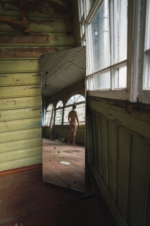 Mirror image of naked man standing at window of abandoned house - KKAF02446