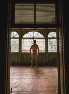 Back view of naked man standing at window of abandoned house - KKAF02447
