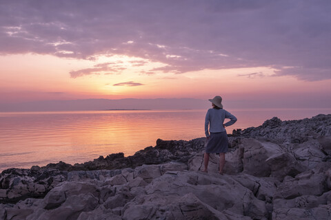 Kroatien, Istrien, Losinj, Frau steht bei Sonnenuntergang an der Felsenküste, lizenzfreies Stockfoto