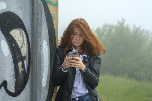 Italy, Finale Ligure, portrait of red headed teenage girl leaning against mural looking at cell phone - LBF02127