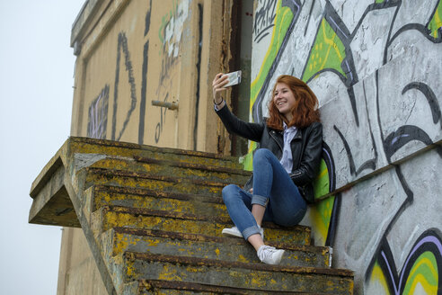 Italy, Finale Ligure, redheaded teenage girl ltaking selfie in front of graffiti wall - LBF02128