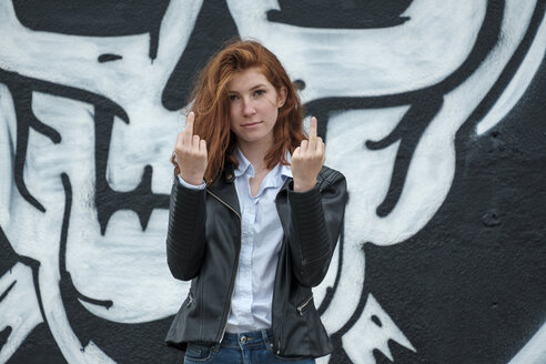 Italy, Finale Ligure, portrait of redheaded teenage girl showing the finger in front of wall with skull graffito - LBF02129