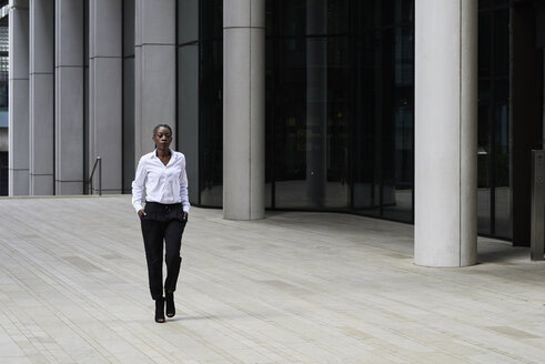Businesswoman wearing white shirt and black trousers walking in front of a modern office building - IGGF00688