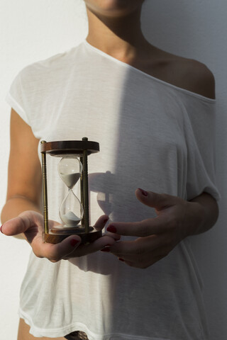 Junge Frau hält eine Sanduhr bei Sonnenuntergang vor einer weißen Wand, lizenzfreies Stockfoto