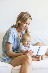 Mother and baby girl sitting on couch looking at picture book - TCF05873