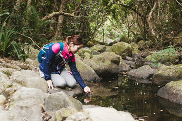 Frau trinkt Wasser aus einem See, während sie auf einem Felsen im Wald sitzt - CAVF49668