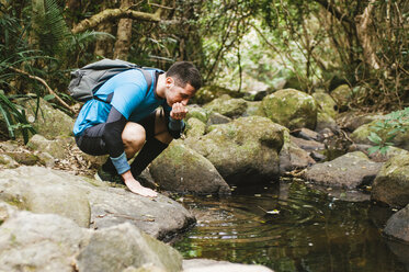 Male hiker drinking water from lake while crouching on rocks in forest - CAVF49669