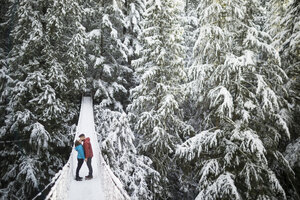 Full length of couple kissing while standing on footbridge amidst trees in forest at Lynn Canyon Park during winter - CAVF49681