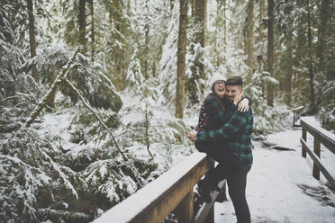 Laughing woman embracing man while sitting on fence in forest at Lynn Canyon Park during winter - CAVF49685