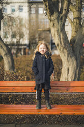 Full length portrait of girl with hands in coat's pockets winking while standing on bench at park - CAVF49700