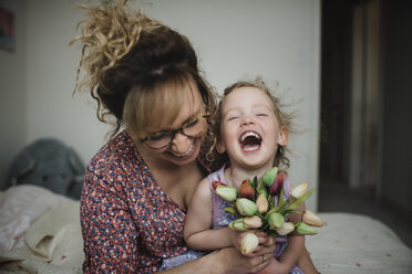 Glückliche Mutter mit Tochter, die Tulpen hält, während sie zu Hause auf dem Bett sitzen - CAVF49716
