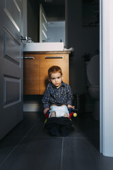 Porträt eines Jungen, der auf dem Töpfchen sitzend seinen Darm entleert, gesehen durch die Türöffnung zu Hause - CAVF49737