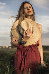 Niedriger Blickwinkel von nachdenklichen Frau hält Hut, während stehend auf grasbewachsenen Feld gegen Himmel - CAVF49745