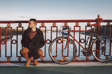Mann schaut weg, während er auf einer Brücke über einen Fluss gegen den Himmel in der Stadt auf einem Fahrrad hockt - CAVF49752