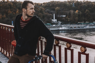 Thoughtful man looking away while standing with bicycle on bridge over river against trees during sunset - CAVF49753