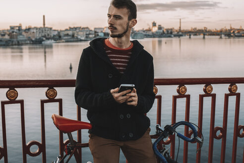 Radfahrer mit Smartphone schaut weg, während er mit dem Fahrrad auf einer Brücke über einen Fluss in der Stadt bei Sonnenuntergang steht - CAVF49754