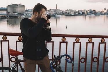 Cyclist photographing with camera while standing with bicycle on bridge over river in city during sunset - CAVF49755