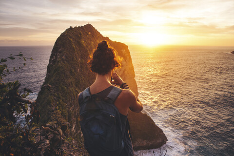 Rückansicht eines Mannes mit Rucksack, der auf einem Berg steht und bei Sonnenuntergang auf das Meer blickt, lizenzfreies Stockfoto