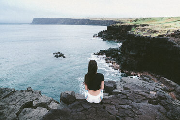 Rear view of woman sitting on rocks at beach against sky - CAVF49783