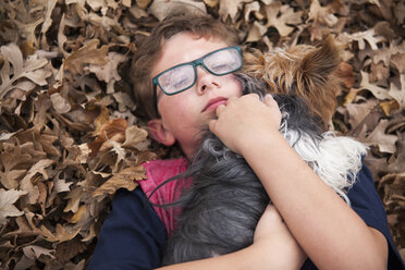 Hoher Blickwinkel des Jungen, der seinen Hund umarmt, während er auf Herbstblättern im Park liegt - CAVF49805